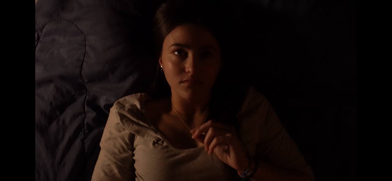 Young woman lying down on a dark bed, softly illuminated with a serious expression.