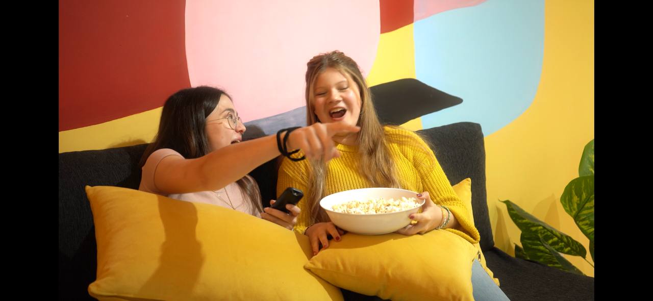 Two young women sitting on a couch with yellow cushions, one holding a bowl of popcorn while the other points excitedly.