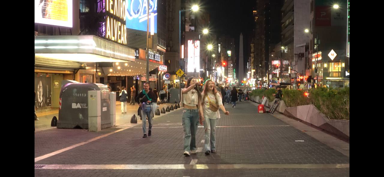 Two young women walking and laughing on a well-lit city street at night with an obelisk monument in the background.