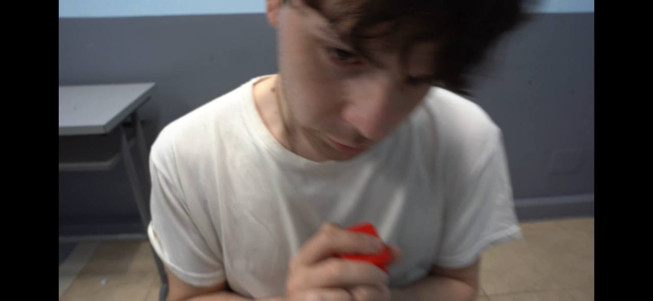 A young person in a white T-shirt holding a red object, with a blurred face and gray walls in the background.