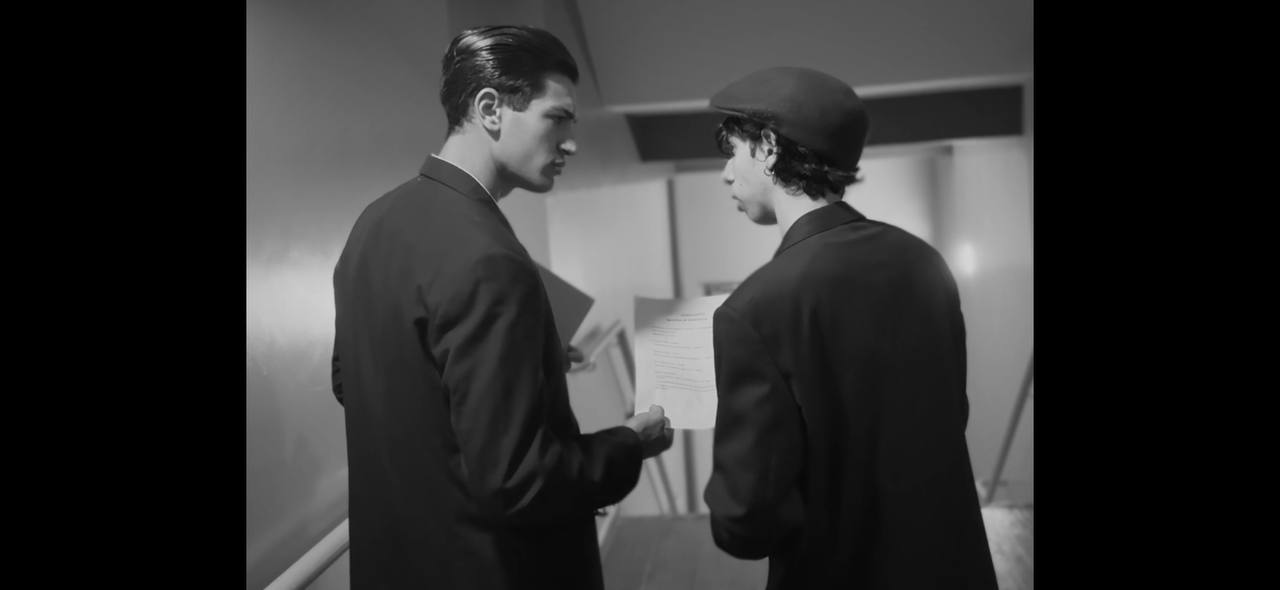 Two men in suits, one wearing a beret, are standing indoors discussing a document.